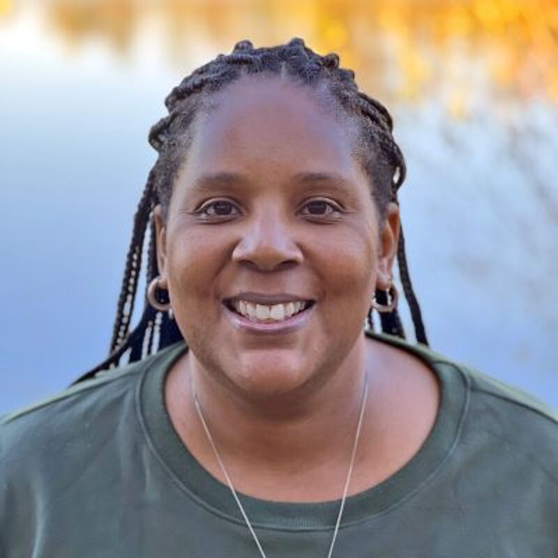 The image shows a Black woman with braided hair, wearing a green shirt and hoop earrings. She is smiling at the camera. The background is blurred, but it appears to be a body of water with trees in the distance. The lighting suggests it is daytime.